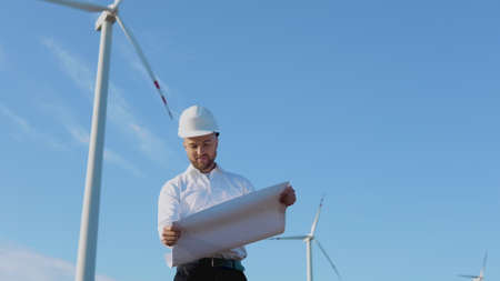 A male engineer in a white helmet and a classic shirt inspects the power plants capacity and reads project drawings on a large sheet of paperの写真素材