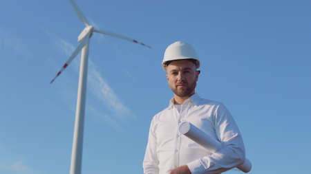 A male engineer in a white helmet and a classic shirt stands near the wind turbines witha large rolled drawings of the project in his handの写真素材