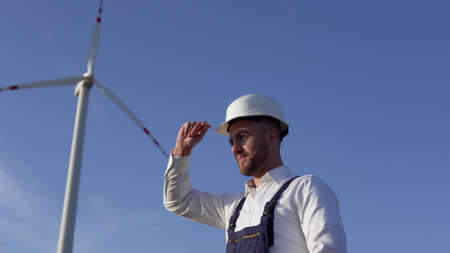 The power plant engineer is standing in a white classic shirt, blue overalls and a white helmet on a windmill backgroundの写真素材