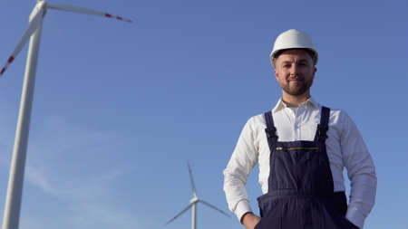The power plant engineer is standing in a white classic shirt, blue overalls and a white helmet on a windmill backgroundの写真素材