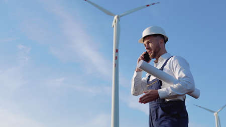 A male engineer in a white helmet, work blue overalls and a classic shirt holds in his hand a large rolled sheet of paper with drawings of the project and talks on the cellphone near the wind turbinesの写真素材