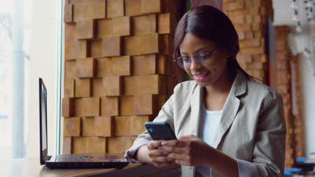 Young indian woman in glasses using phone while sitting in cafe with laptopの写真素材