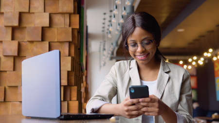 Young indian woman in glasses using phone while sitting in cafe with laptopの写真素材