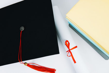 A black square graduate hat with a red tassel lies on a diploma and a stack of books. Education...