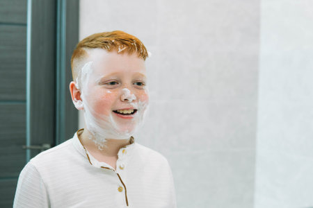 Funny boy in the bath smears his face with shaving cream, the child imitates an adult dad shaves. The boy smeared his cheeks with shaving foamの写真素材