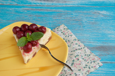 Yellow plate, mint leaf, a piece of cheesecake with cherries and metal fork with napkin on blue wooden shabby background. Copy space for textの写真素材