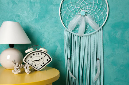White mint dream catcher, alarm clock and table lamp on yellow table in bedroom interior on aquamarine background. Copy space for text.の写真素材