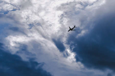 airplane silhouette flying on dark blue and white cloudy background overcast awayの写真素材
