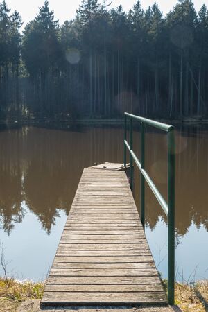 A wooden footbridge on the lake, also used as a summer boat dock. The forest behind the lake, reflections in the calm water.の写真素材