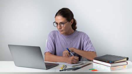 A woman with a laptop, books, notebooks, notebooks at the table makes notes - online learning,...