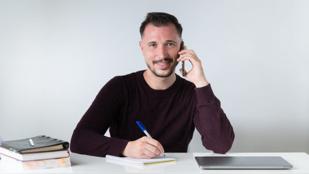 A man talks on the phone and takes notes in a notebook at an office desk.
