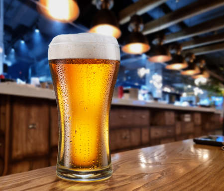 Cooled glass of beer with condensate drops on the wooden table. Blurred pub interior at the background.の写真素材
