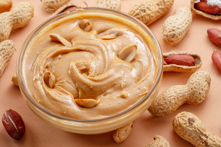 Bowl of peanut butter and peanuts around it on beige background. top view.