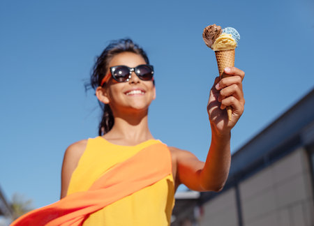 Happy, smiling girl holding ice cream cone with colorful ice cream balls. Sunny sea coastline at the background.の写真素材
