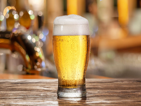 Glass of chilled beer on wooden bar table top and blurred bar interior at the background.の写真素材