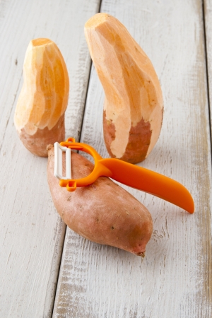 Three  sweet potatoes, two are partially peeled, and vegetable peeler on wooden table の写真素材