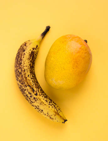 Over ripe banana with dark spots and mango fruits on yellow background.の写真素材