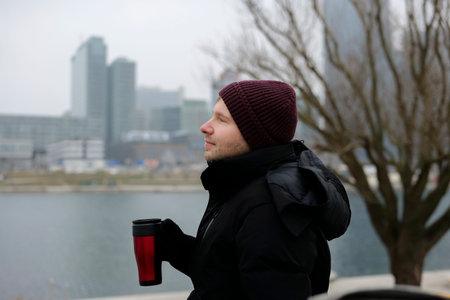 Young man enjoying a warm drink from a disposable cup near the river on a chilly day in winter