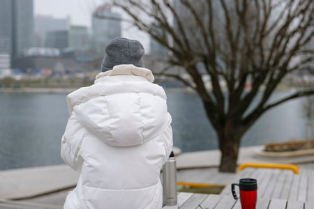 Young woman in a white puffer jacket sits on a bench, overlooking a foggy city waterfront. Concept of an urban winter activityの写真素材