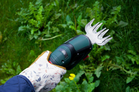 Womans hand holds a green electric grass shears among grass, weeds and small wildflowers in garden