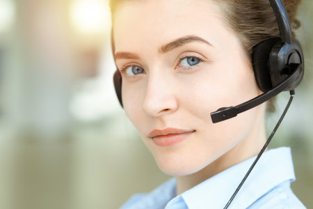 Beautiful female specialist with headphones helping customers online and smiling charmingly in sunny call center office. Business people conceptの写真素材