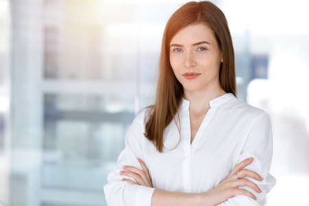 Beautiful female specialist with arms crossed standing in sunny office and smiling charmingly. Business people conceptの写真素材