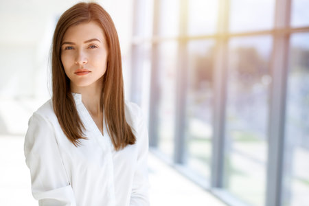 Beautiful female specialist with dark hair standing in sunny office and smiling charmingly. Working...