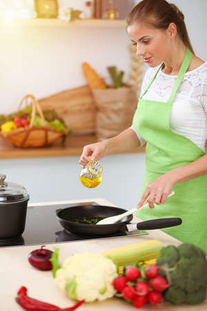 An attractive young dark-haired woman frying meal at worm pan by new keto recipe while standing and smiling in sunny kitchen. Cooking and householding conceptsの写真素材