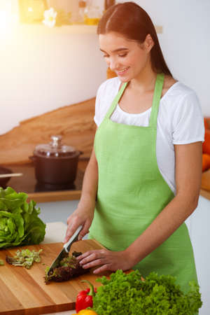An attractive young dark-haired woman preparing delicious fresh vegetarian salad while standing and smiling in sunny kitchen. Cooking and householding conceptsの写真素材