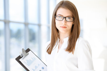Beautiful female specialist with clipboard standing in modern office and smiling charmingly. Working on design, data analysis, plan strategy. Business people conceptの写真素材