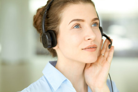 Beautiful female specialist with headphones helping customers online and smiling charmingly in modern call center office. Business people conceptの写真素材