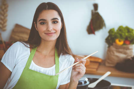 An attractive young dark-haired woman tasting ready hot meal with a wooden spoon while standing and smiling near the kitchen stove. Cooking and householding conceptsの写真素材