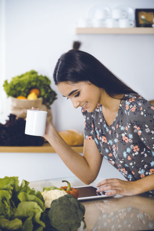 An attractive young dark-haired woman drinking morning coffee and looking for a new recipe for a delicious meal while sitting at the kitchen table. Tablet pc is the best cookbookの写真素材