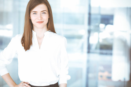 Beautiful female specialist with dark hair standing in modern office and smiling charmingly. Working...
