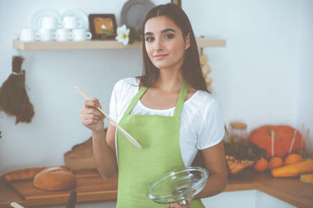 An attractive young dark-haired woman tasting ready hot meal with a wooden spoon while standing and smiling near the kitchen stove. Cooking and householding conceptsの写真素材