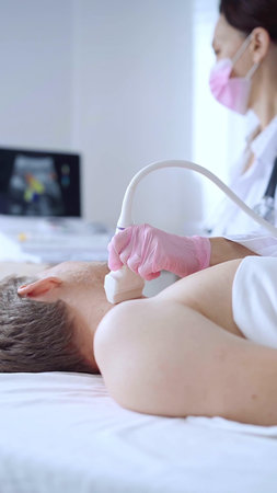 Doctor woman with pink medical gloves and mask performing neck ultrasound exam on male patient....