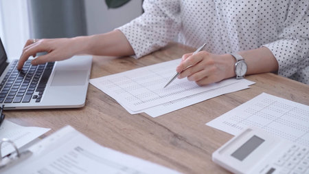 Close-up of a womans hand checking financial paper beside a laptop on the wooden desk. Bookkeeping,...