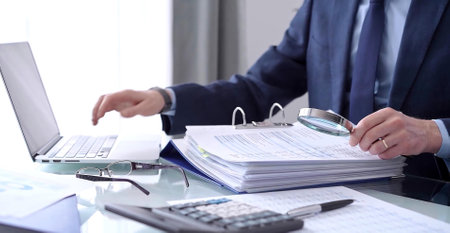 Businessman or auditor is looking through binders with papers while using a calculator and magnifying glass for financial documents analyzing. Audit and taxes conceptの写真素材