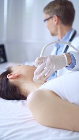 Doctor man wearing white protective gloves is using ultrasound equipment on the neck of patient woman, lying for examination. Medicine conceptの写真素材