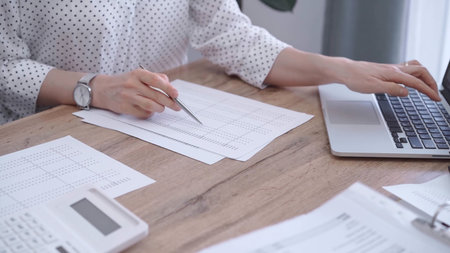 Close-up of a womans hand checking financial paper beside a laptop on the wooden desk. Bookkeeping, finance audit and taxesの写真素材