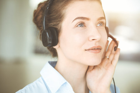 Beautiful female specialist with headphones helping customers online and smiling charmingly in sunny...