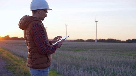 Man engineer, wearing a white protective helmet is taking notes with a tablet computer in a field with wind turbines, as the sun sets. Clean energy and engineering conceptの写真素材