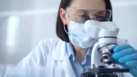 Female scientist in lab goggles, protective mask and gloves is using microscope in laboratory....