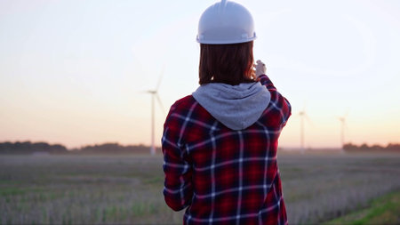 Woman engineer taking notes on a clipboard on a field with wind turbines, as the sun sets, back view. Clean energy and engineering conceptの写真素材
