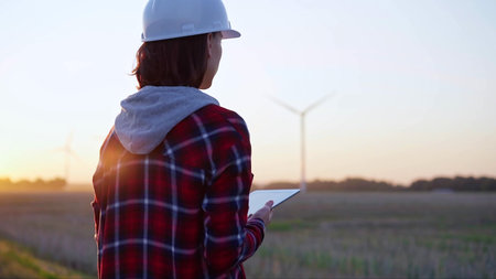 Woman engineer taking notes with a tablet computer on a field with wind turbines, as the sun sets. Concept of clean energy and engineeringの写真素材