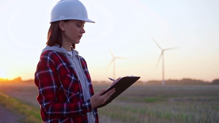 Female engineer taking notes on a clipboard on a field with wind turbines, as the sun sets. Concept...