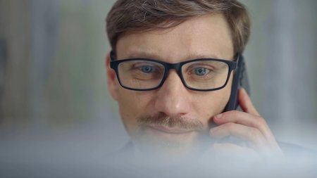 Businessman with glasses speaking on the phone, partially obscured by blurred laptop. Front view and...