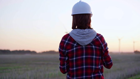 Woman engineer taking notes on a clipboard on a field with wind turbines, as the sun sets, back...