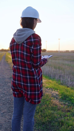 Woman engineer wearing a white protective helmet is taking notes with a clipboard in a field with...