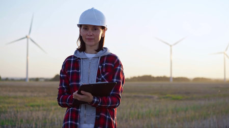 Female engineer is standing straight and keeping clipboard on a field with wind turbines, as the sun...